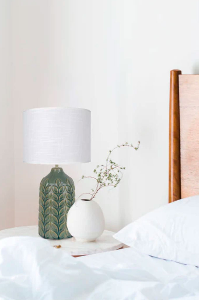 Green ceramic table lamp  with a white linen drum shade next to a bed over a night table