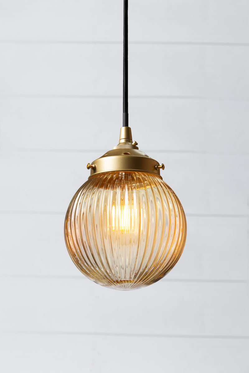 Amber reeded glass ball shade on a black pendent cord with gold/brass metalware. 