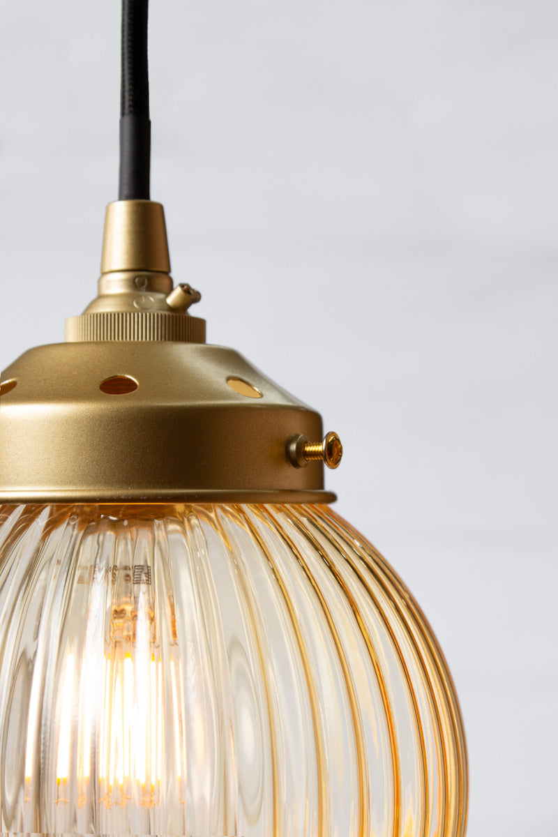 Close up of an amber reeded glass ball shade on a black pendent cord with gold/brass metalware. 