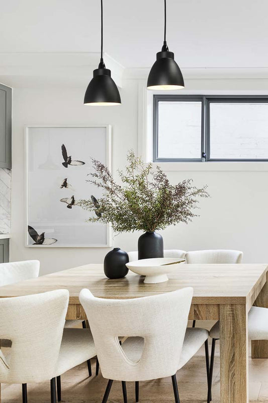 Two black cellar light pendants on black pendant cords hanging in a white dining space. 