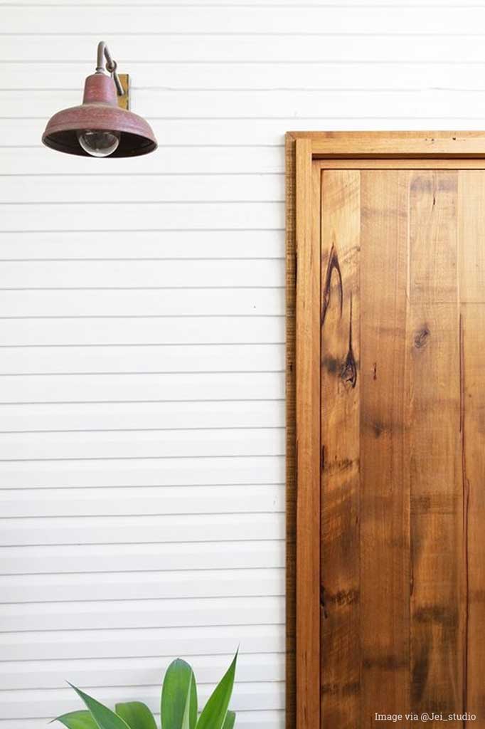 Copper outdoor wall light, mounted on white weather board next to a timber door. 