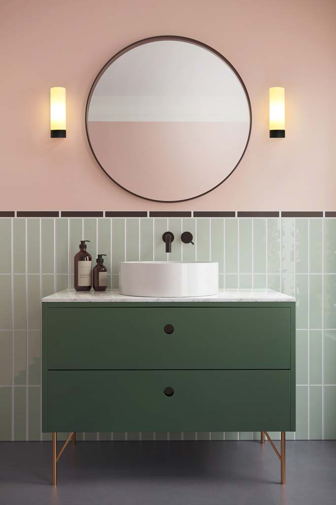 Two simple cylindrical wall sconces with black hardware installed next to a round mirror and green vanity. 