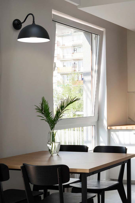 Black dome shade hanging from a gooseneck wall sconce in a a dining space with natural light. 