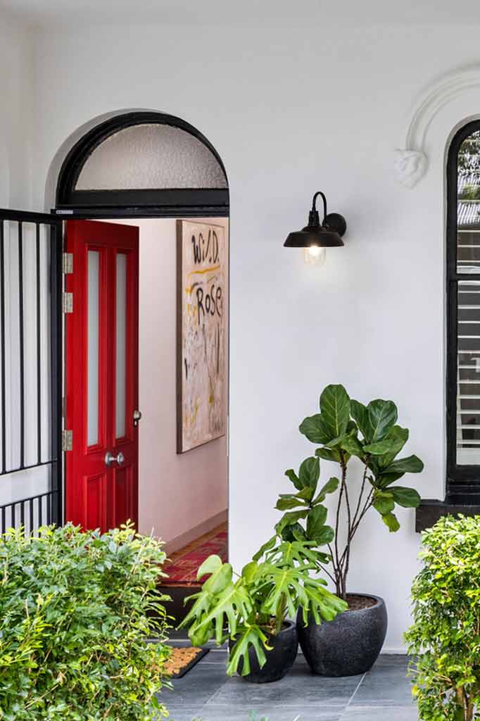 A black Farmington Gooseneck Wall Light shown installed on a front porth with white walls and a bright red door. 