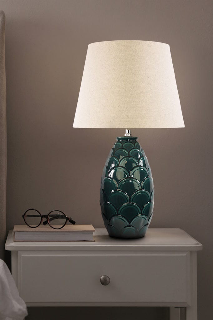 Table lamp with dark green scale-like base and shade in an ivory finish on a bedside table.